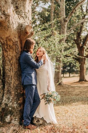 A couple shares an intimate romantic moment against a large tree trunk in a sunlit woodland garden setting.