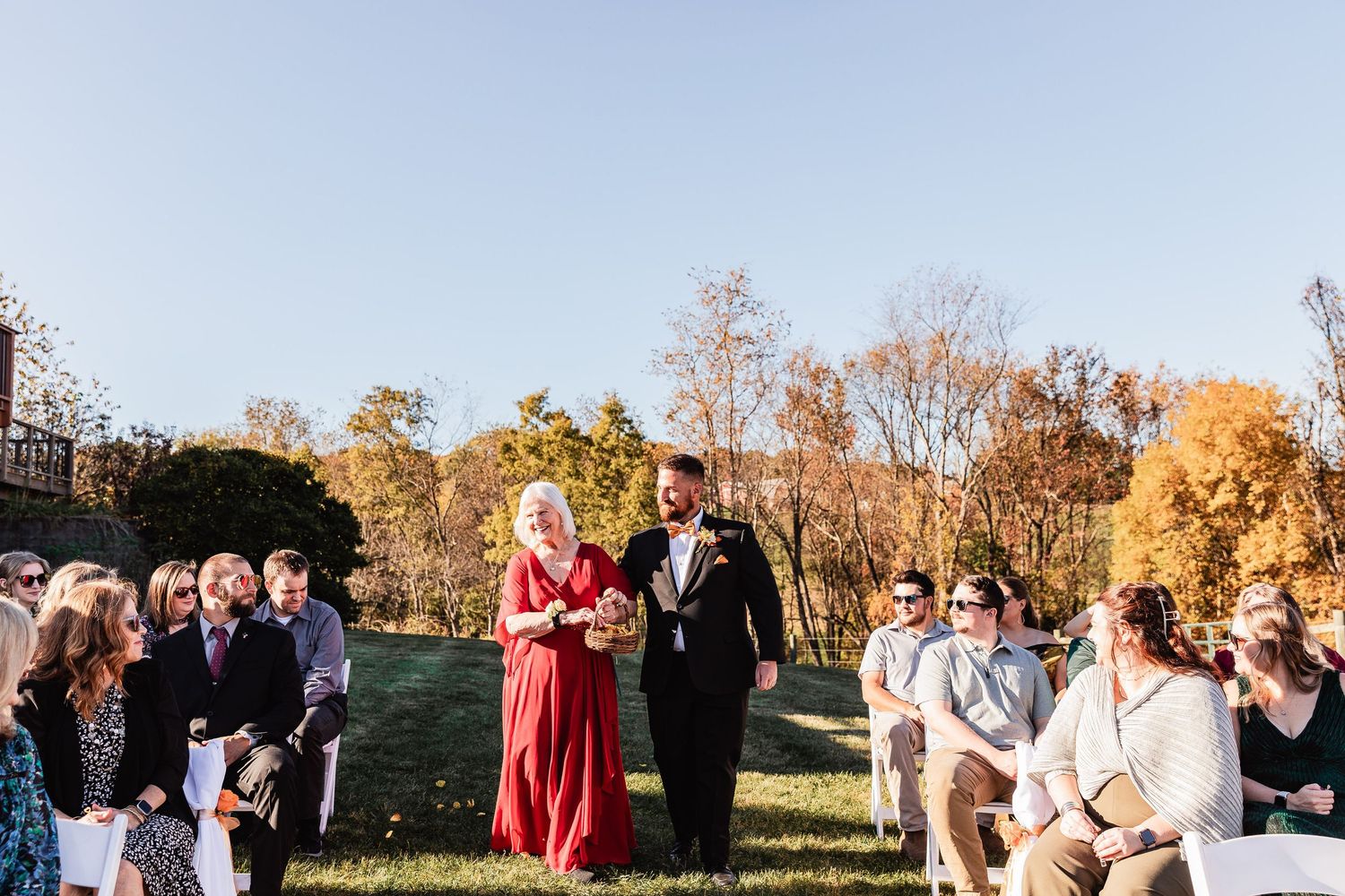An outdoor ceremony in a field with fall foliage, guests seated on chairs and people standing at the front.