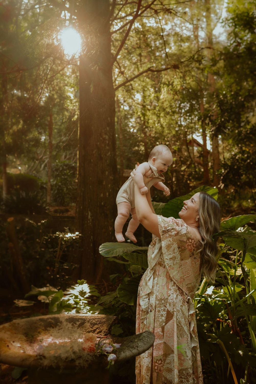 Woman in floral dress lifts baby in sunlit forest setting with golden sunlight streaming through trees.
