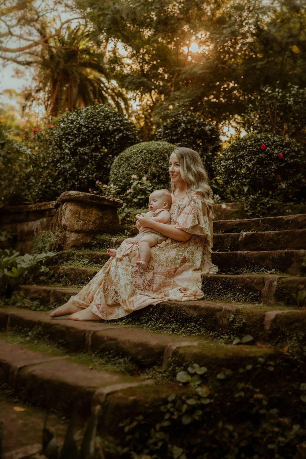 A romantic photo of someone in a flowy floral dress sitting on weathered stone garden steps at golden hour sunset.