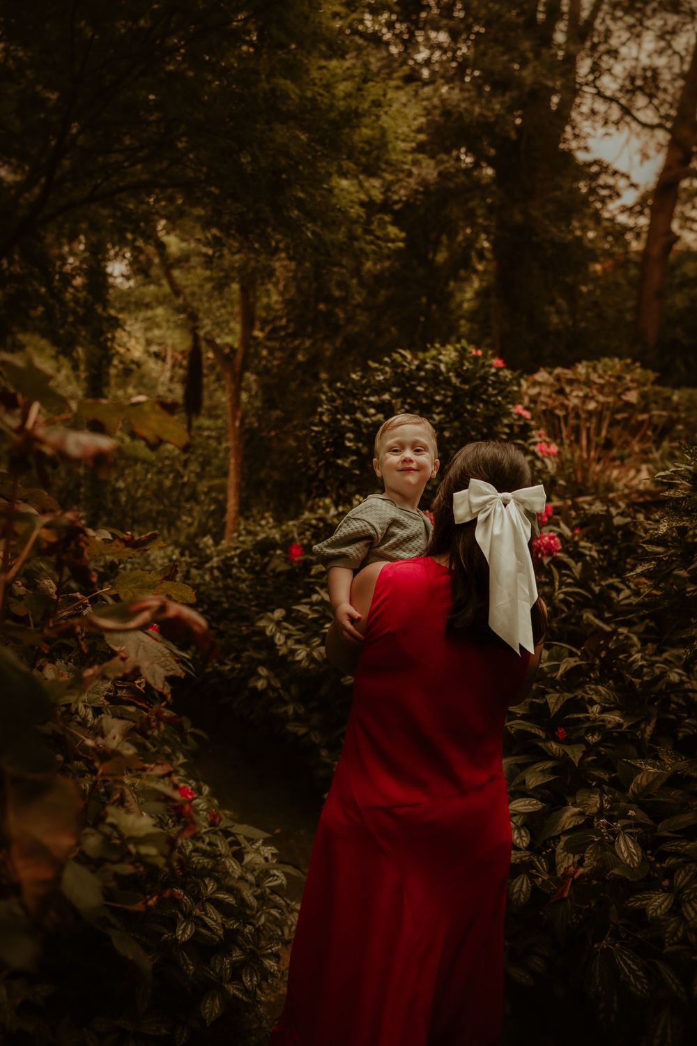 Little one in a red velvet dress twirls in a lush fall garden setting with golden sunlight filtering through trees.