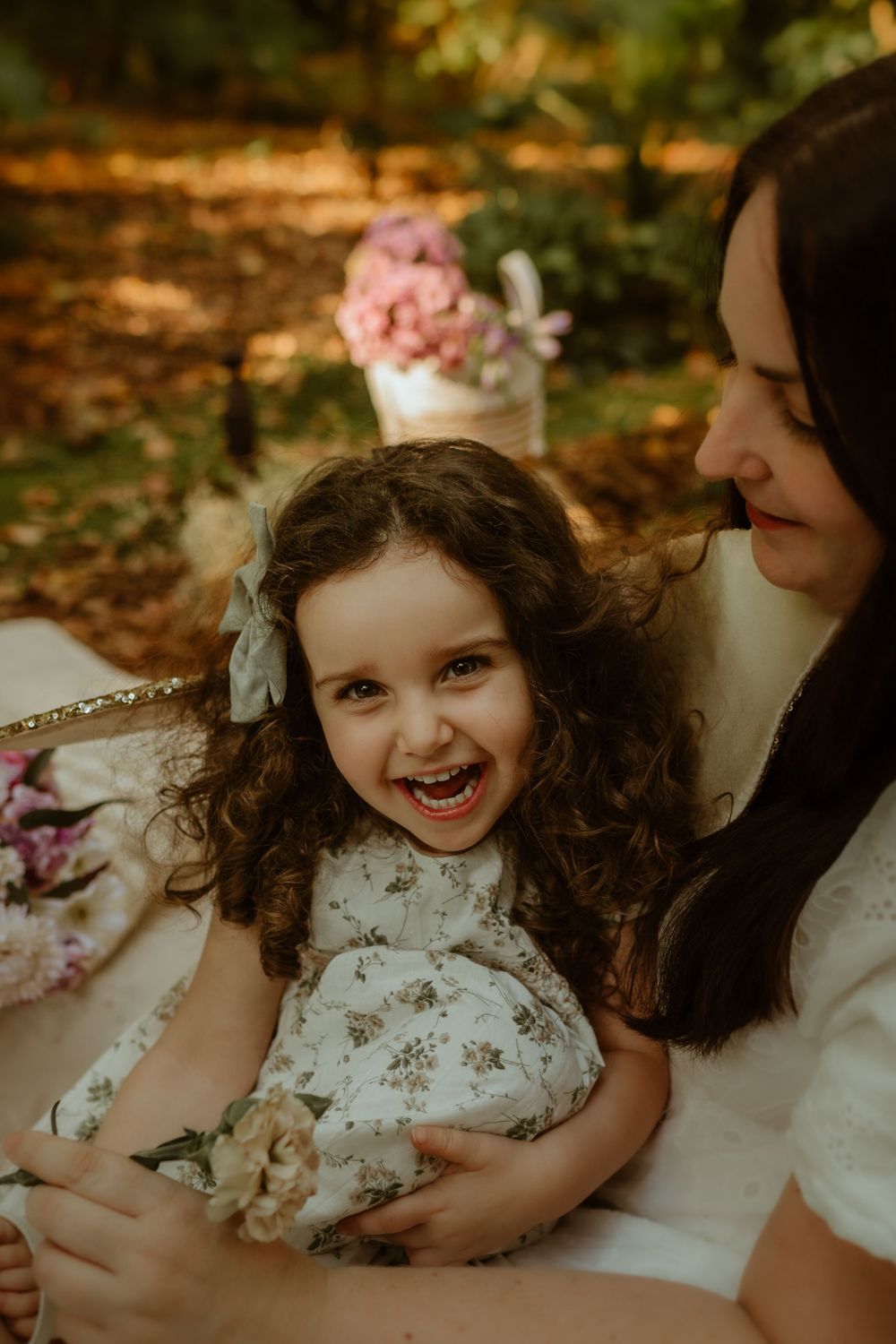 Young girl in white dress laughs joyfully while being held in a warm autumn setting with fallen leaves.