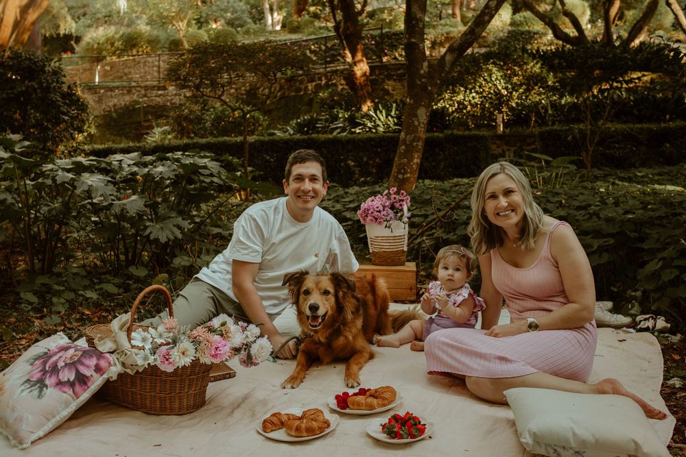 A family enjoys a cozy outdoor picnic on a blanket with their golden retriever dog and decorative baskets in a garden setting.