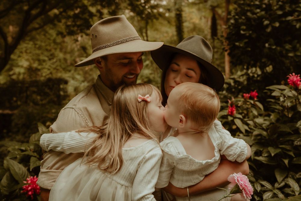 Family wearing beige and white clothing sharing intimate moment together in garden with blooming red flowers.