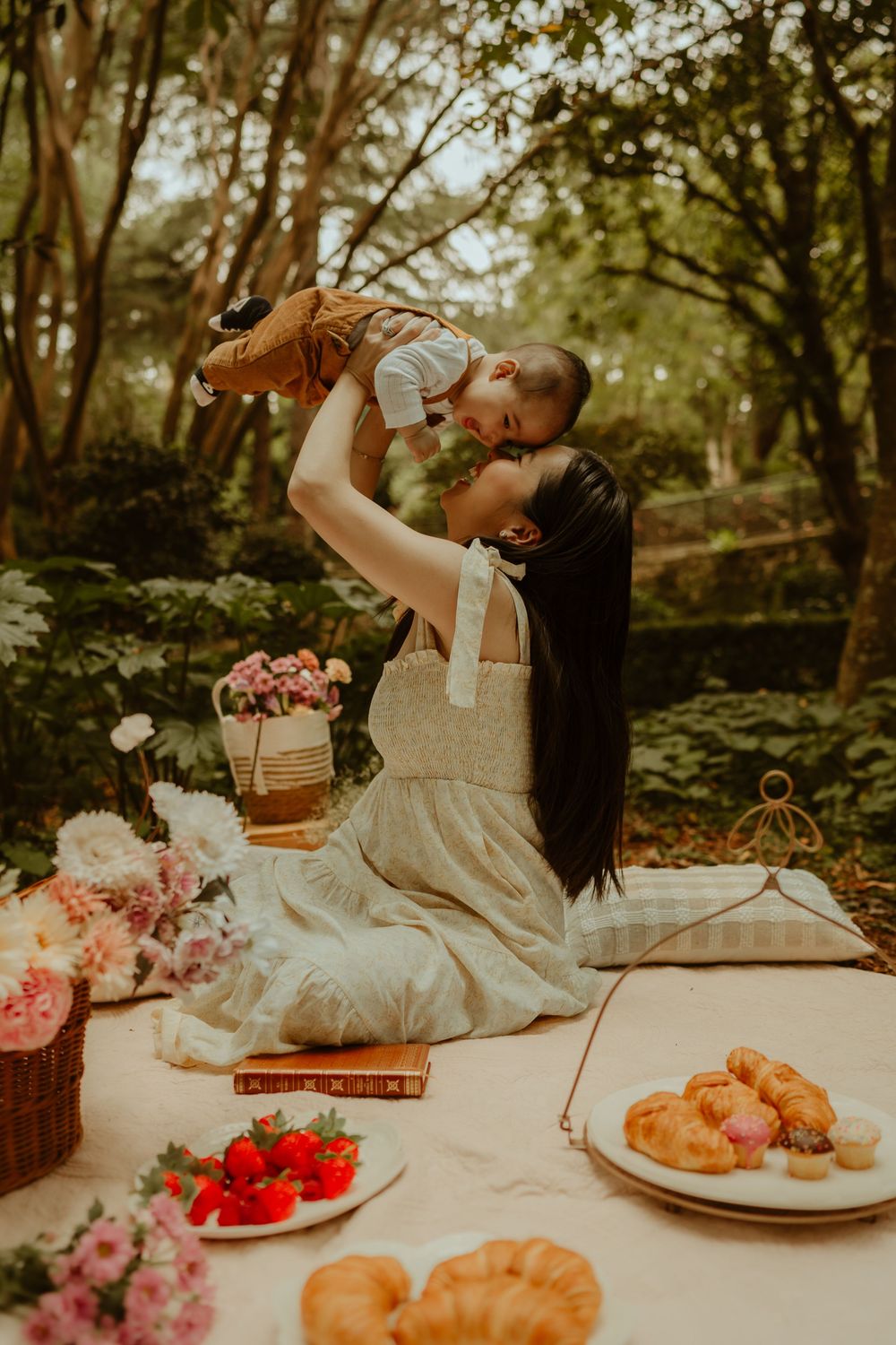 Person in white dress lifts up small child during picnic in forest with flowers and pastries on blanket.