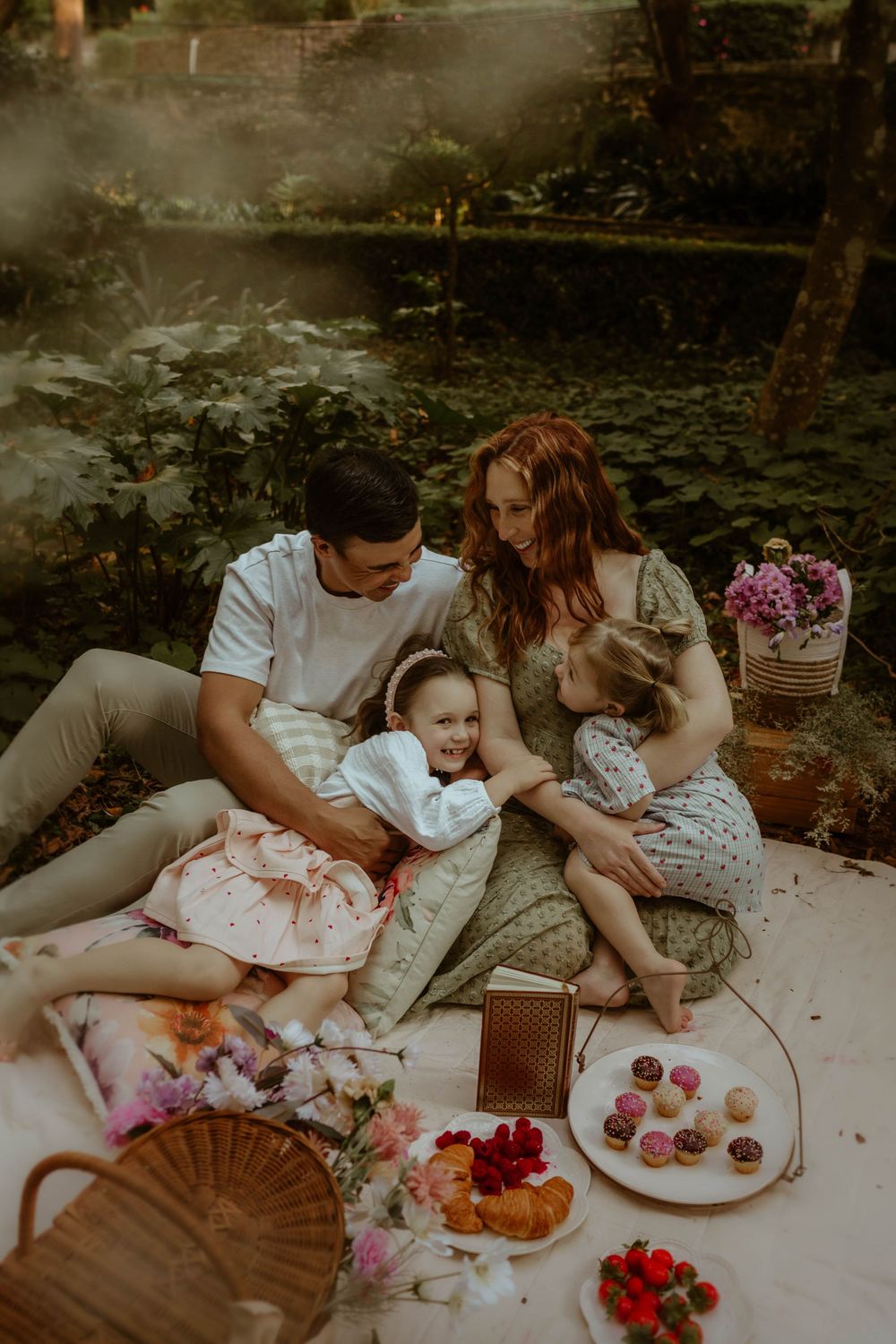 Young family enjoys a romantic outdoor picnic with desserts and flowers on a blanket in a dreamy forest setting.