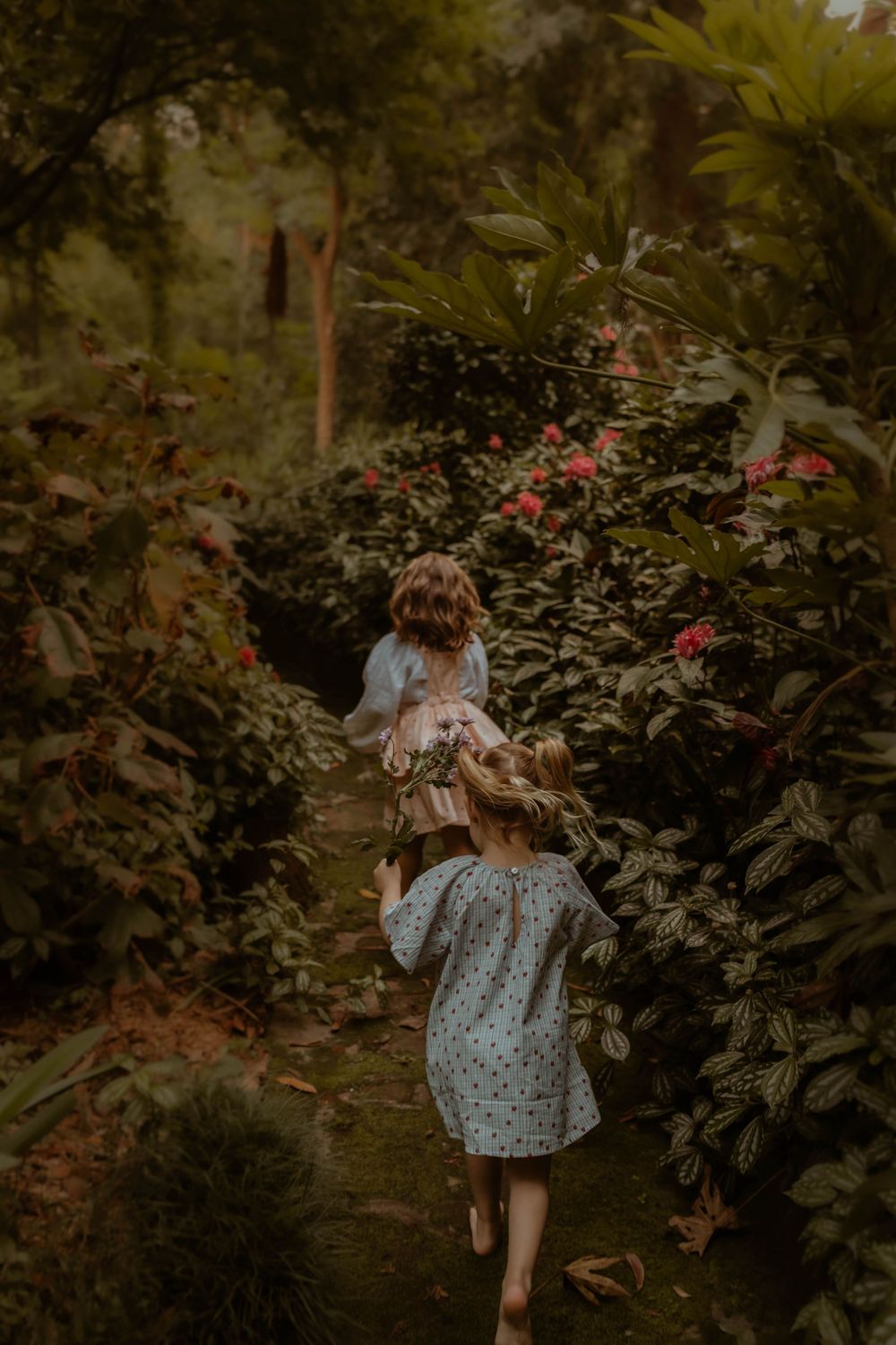 A person wearing a floral dress explores a lush garden with flowering plants and greenery in moody lighting.