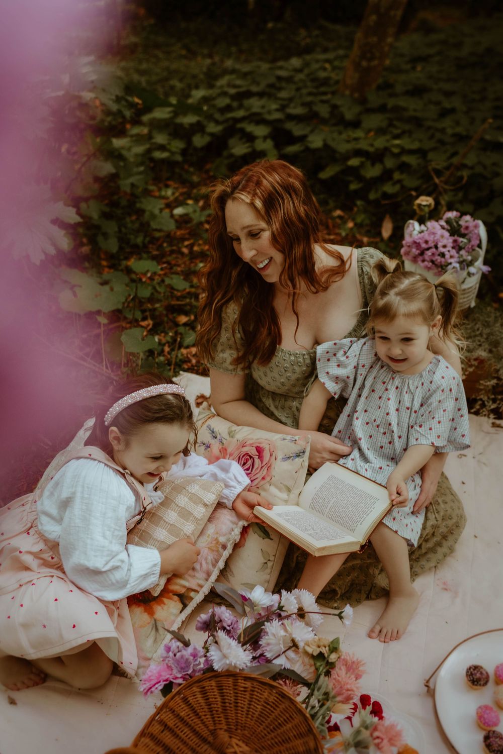 A woman and two young children sharing a picnic blanket outdoors with books and flowers scattered around them.