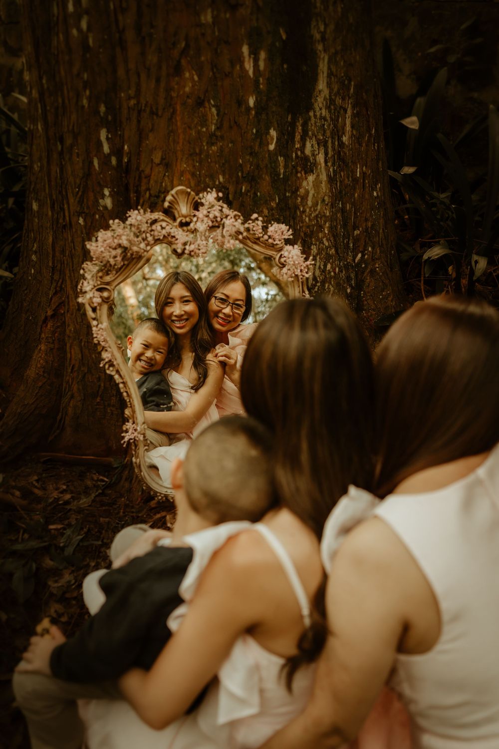 Vintage ornate mirror reflecting standing subjects in white dresses against a rustic tree backdrop in warm tones.
