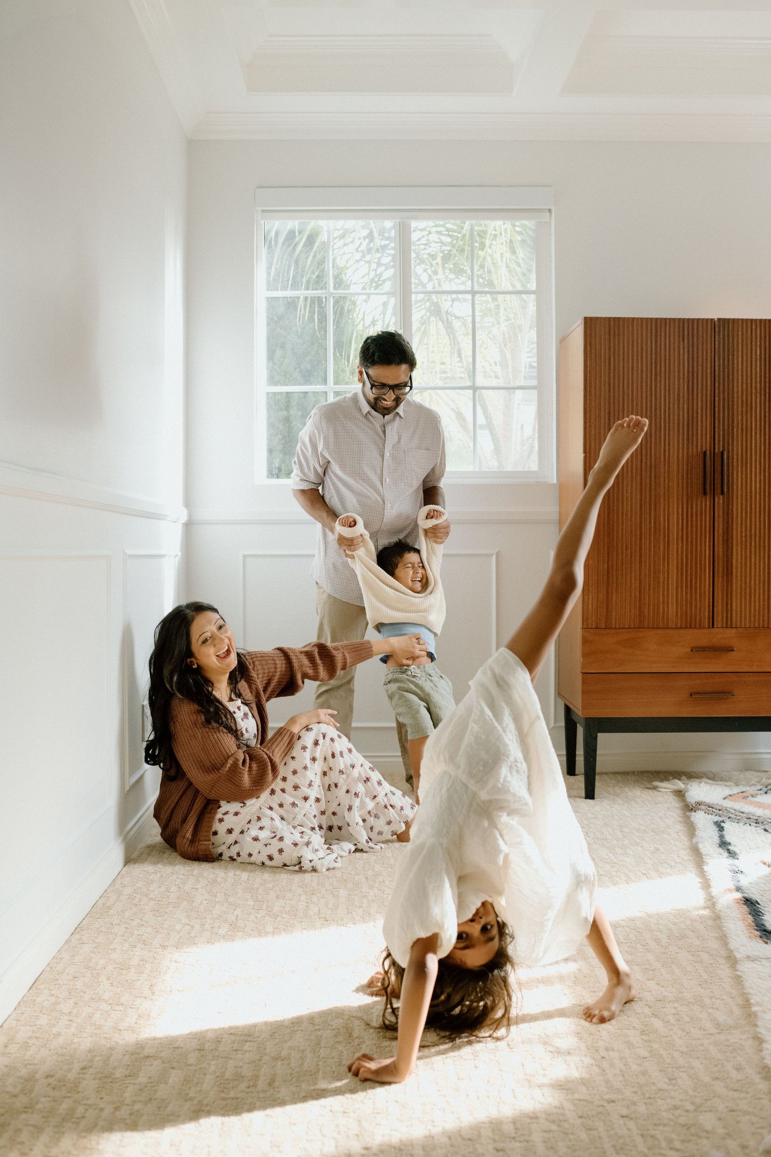 A family plays together in a bright sunlit room with white walls and a wooden wardrobe.