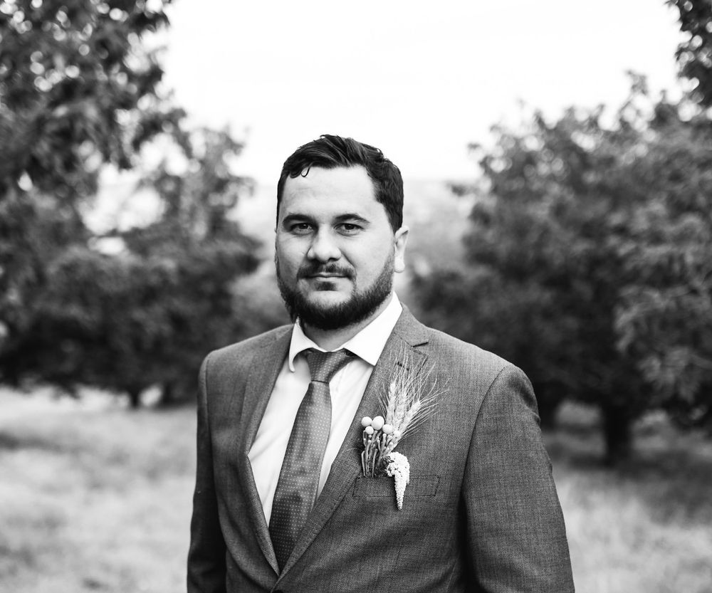 A formal portrait of someone wearing a grey suit with a boutonniere against a natural backdrop.