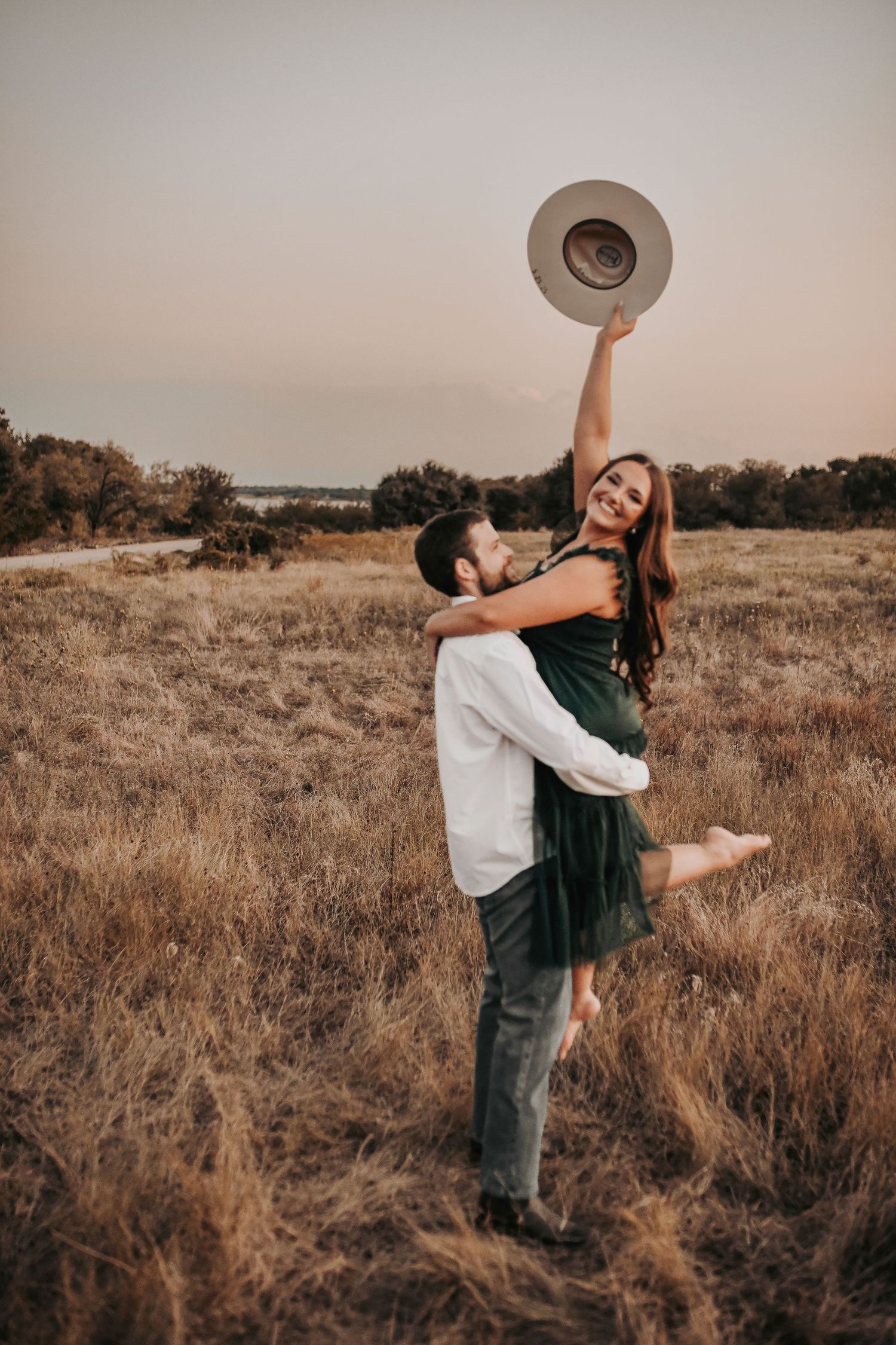 a happy couple laugh as he lifts her and as she lifts her cowgirl hat and smiles at the camera for her sunset engagement photos