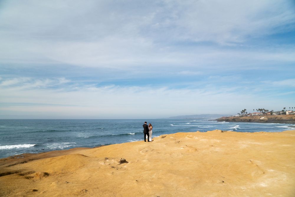 A couple stands on a sandy beach overlook with blue ocean waves and cloudy sky in San Diego, California.