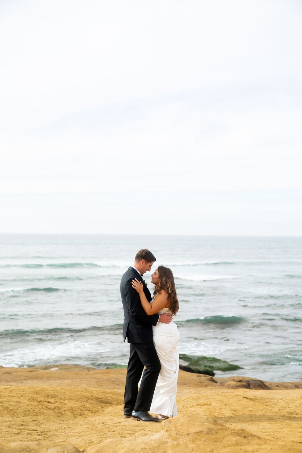 A couple in formal attire embraces romantically on a sandy beach cliff overlooking the ocean at sunset.