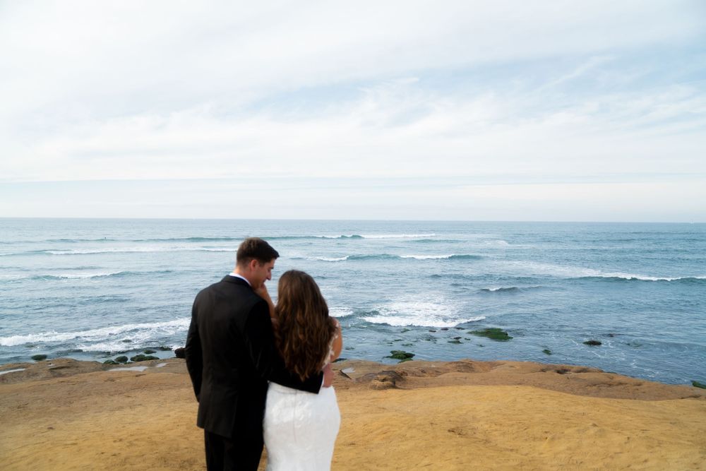 Couple in formal attire embracing while looking out at the ocean from a rocky coastal cliff at sunset.