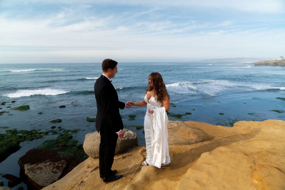 Two people share a romantic moment while standing on rugged coastal cliffs overlooking the Pacific Ocean on a sunny day.