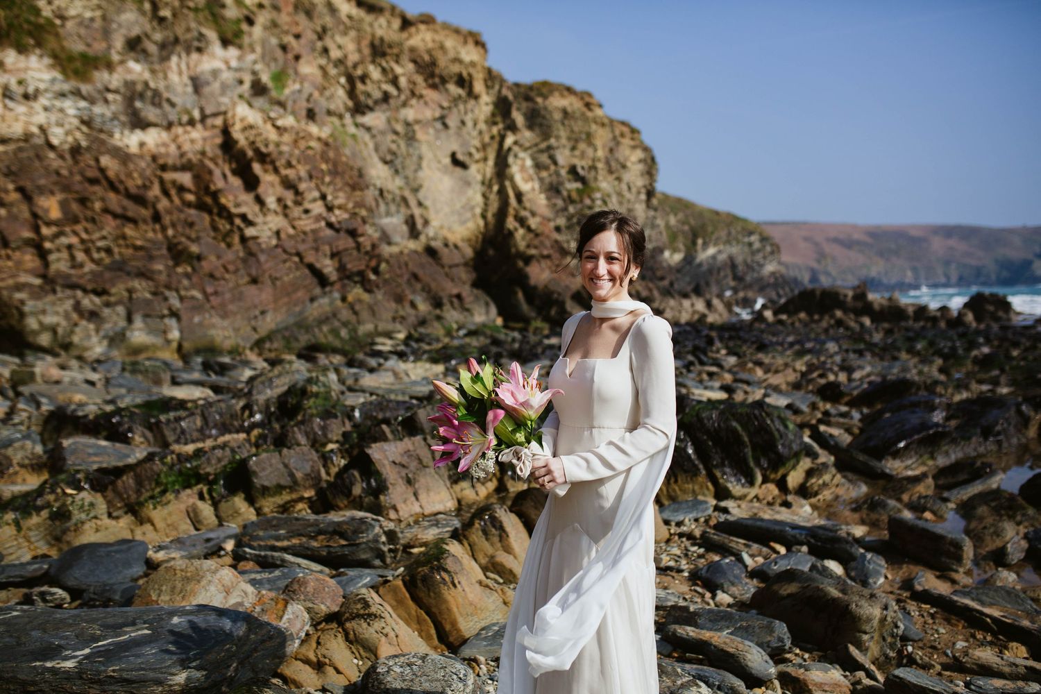Someone in a flowing white wedding dress holds a vibrant pink bouquet while standing on rocky coastal cliffs by the ocean.