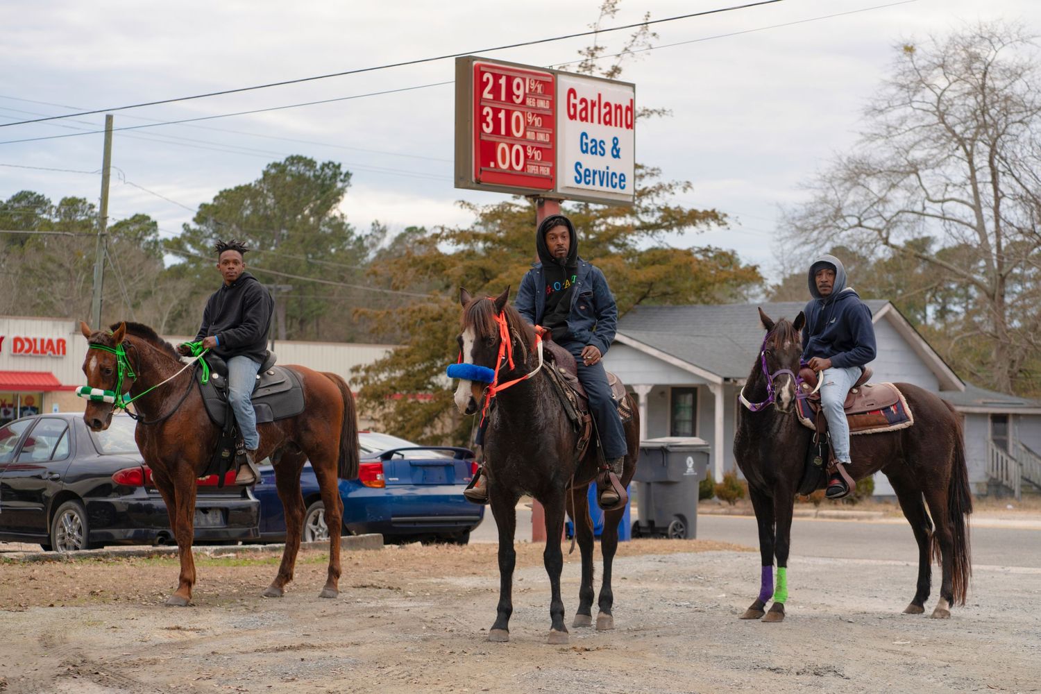 Three horseback riders gather in front of a small business on a rural street during autumn.