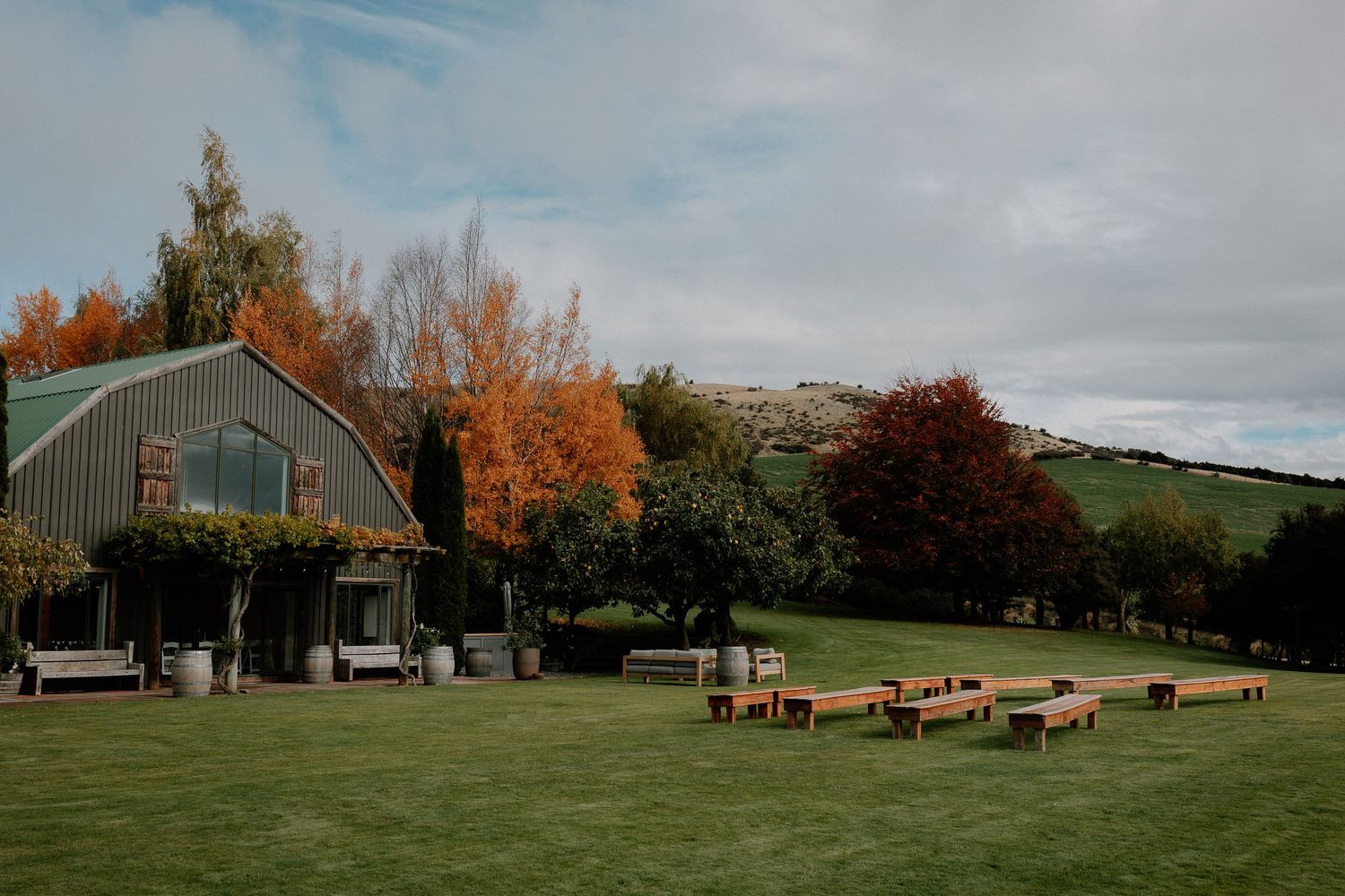 Outdoor wedding venue with wooden benches arranged on a lawn near a green barn surrounded by autumn trees.