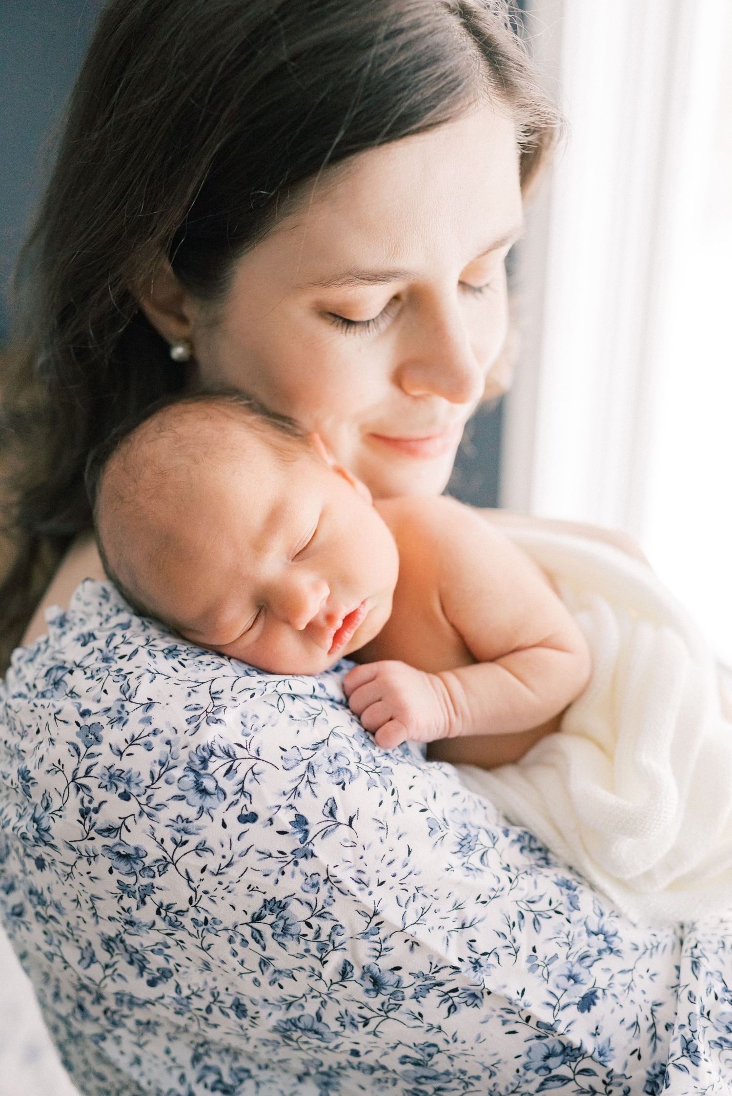 A sleeping newborn nestles against a blue and white floral dress near a window in soft natural light.