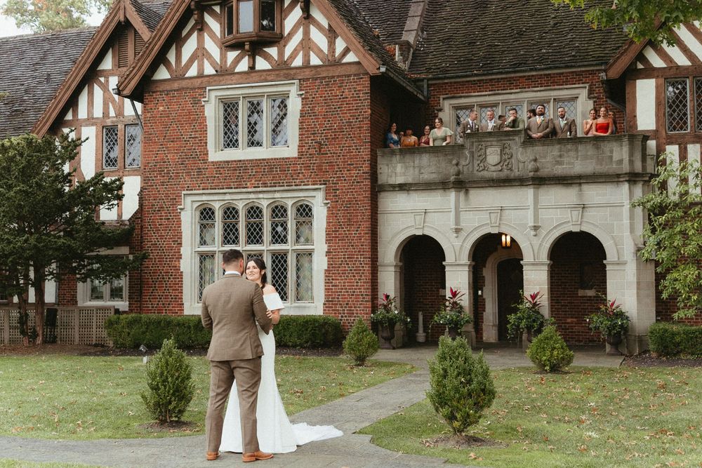 A couple stands outside a Tudor-style brick mansion with arched entryways and ornate architectural details.