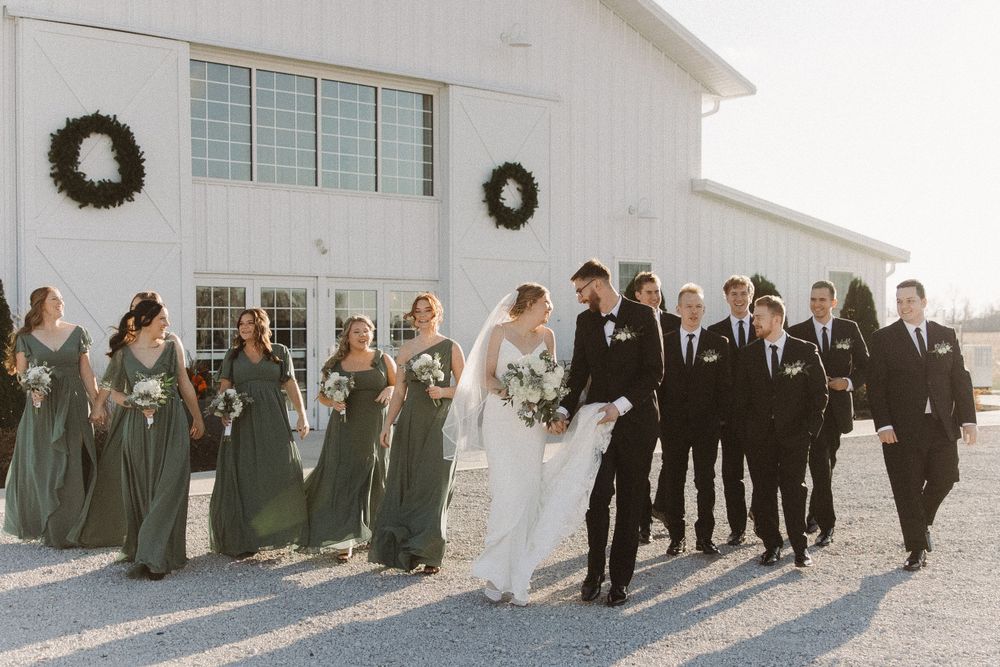 A wedding party in sage green dresses and black suits walks together outside a white church building with wreaths on the wall.