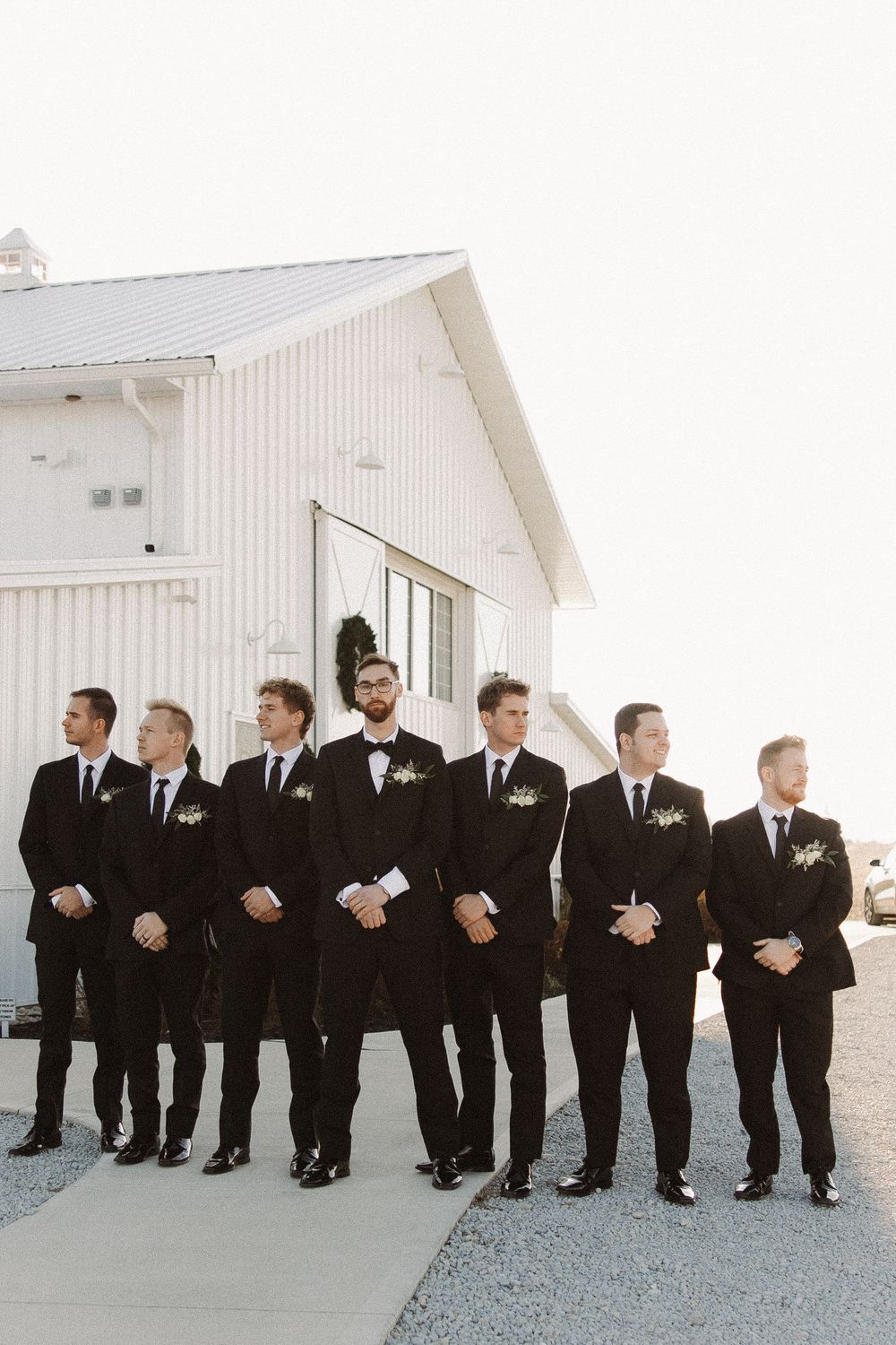 Seven groomsmen in black suits and ties stand in front of a white barn building for a formal wedding photo.