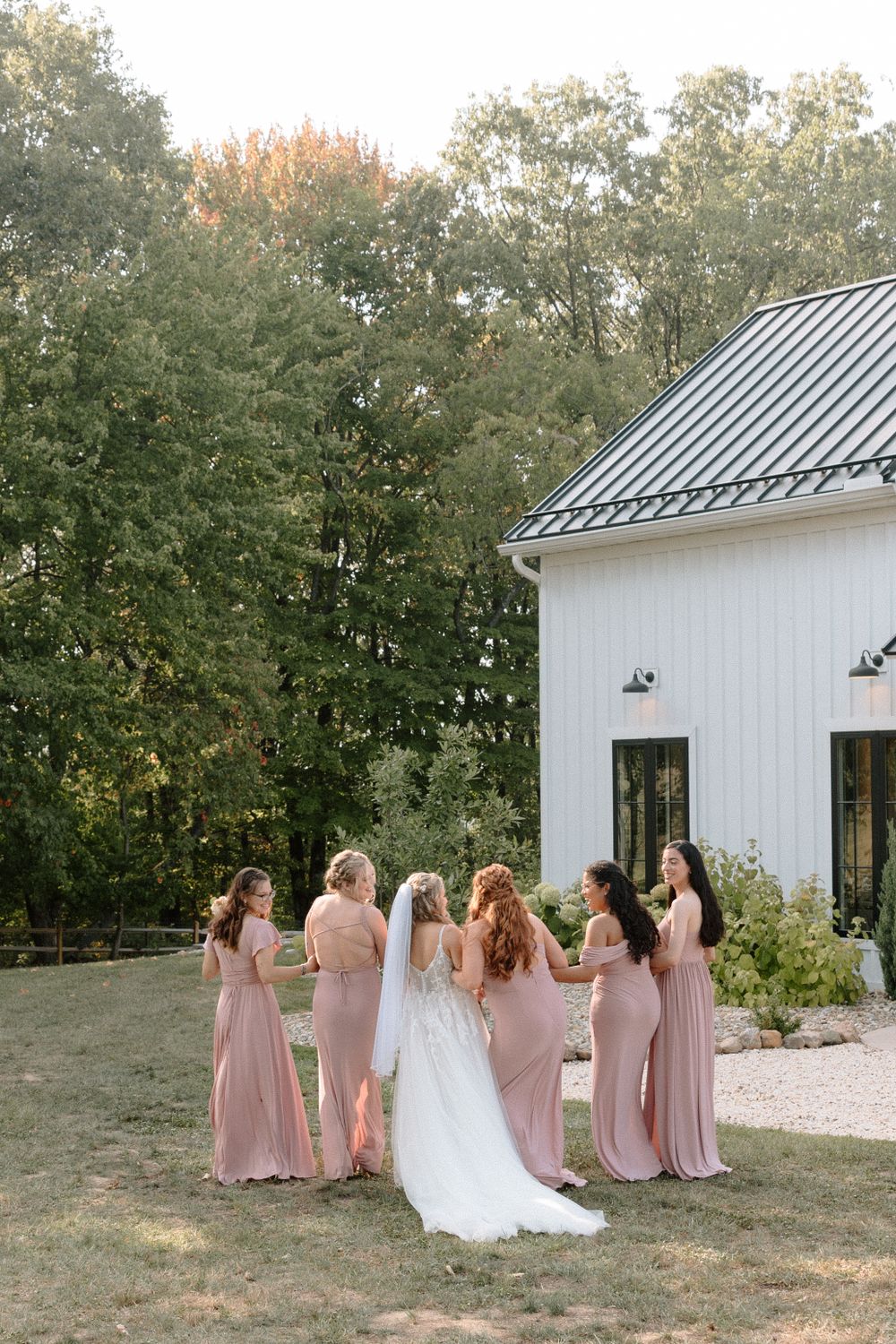 A group in blush pink gowns and a white dress gather outside a modern white barn building surrounded by trees.