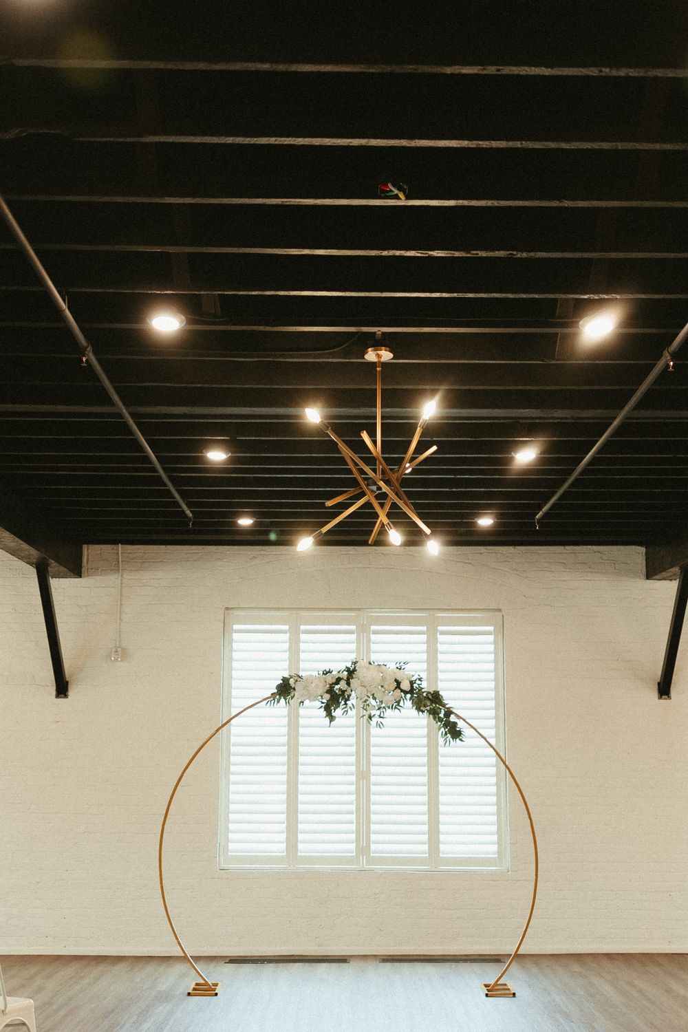 Modern event space with black exposed ceiling beams, gold circular arch and floral arrangement against white shuttered window.