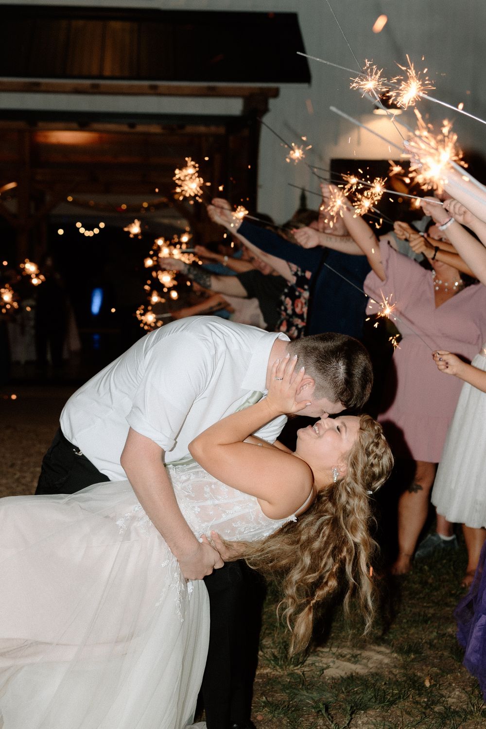 A couple shares a romantic kiss during their wedding sparkler exit as guests hold glowing sparklers overhead.