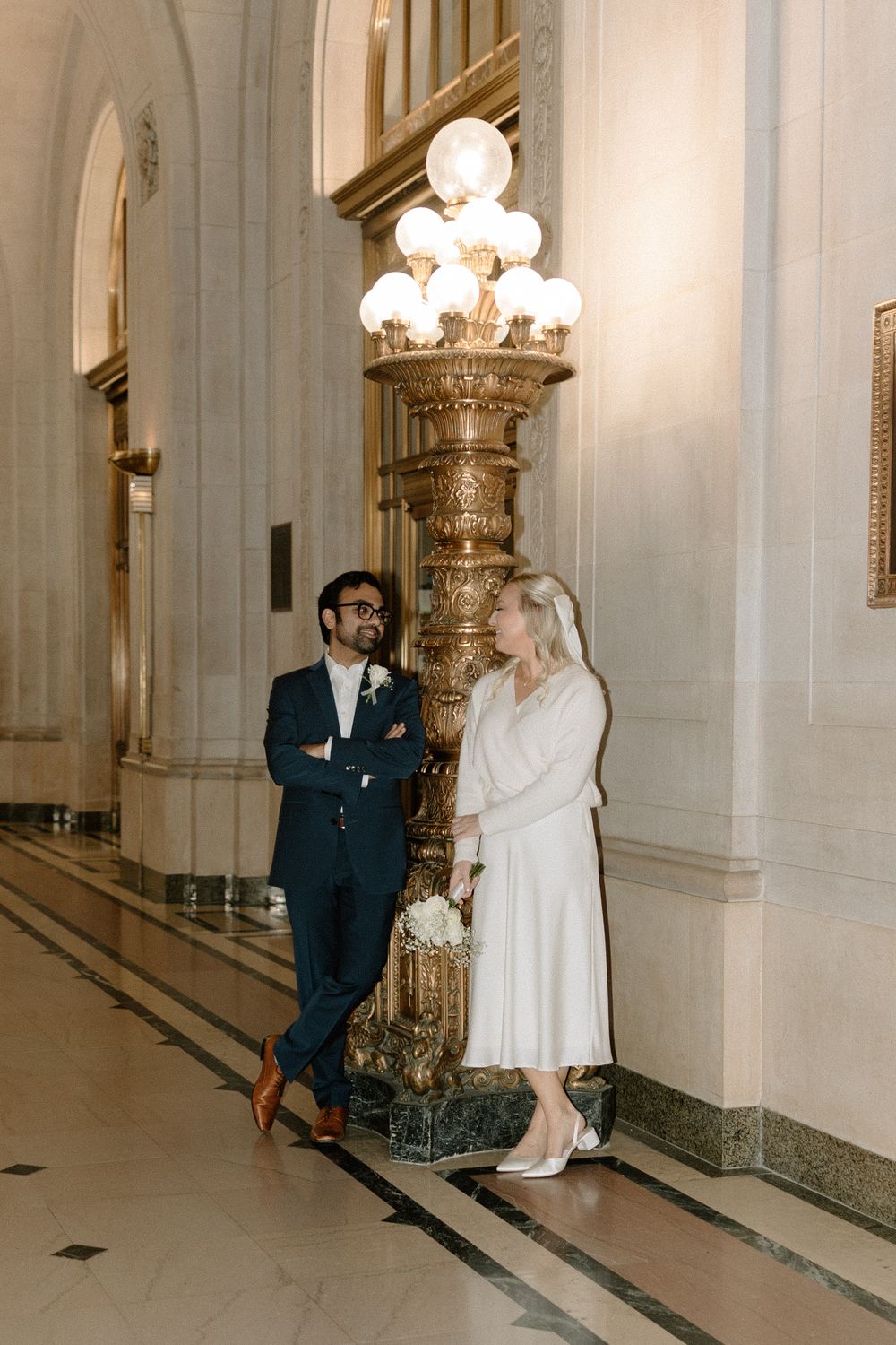 Two people having a conversation beside an ornate vintage lamp post with glowing white globes in an elegant hallway.