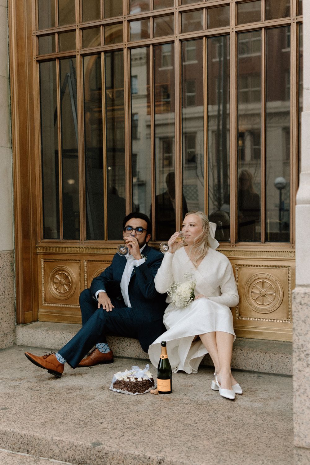 A couple sits on stone steps sharing champagne and snacks outside an ornate building with wooden doors.