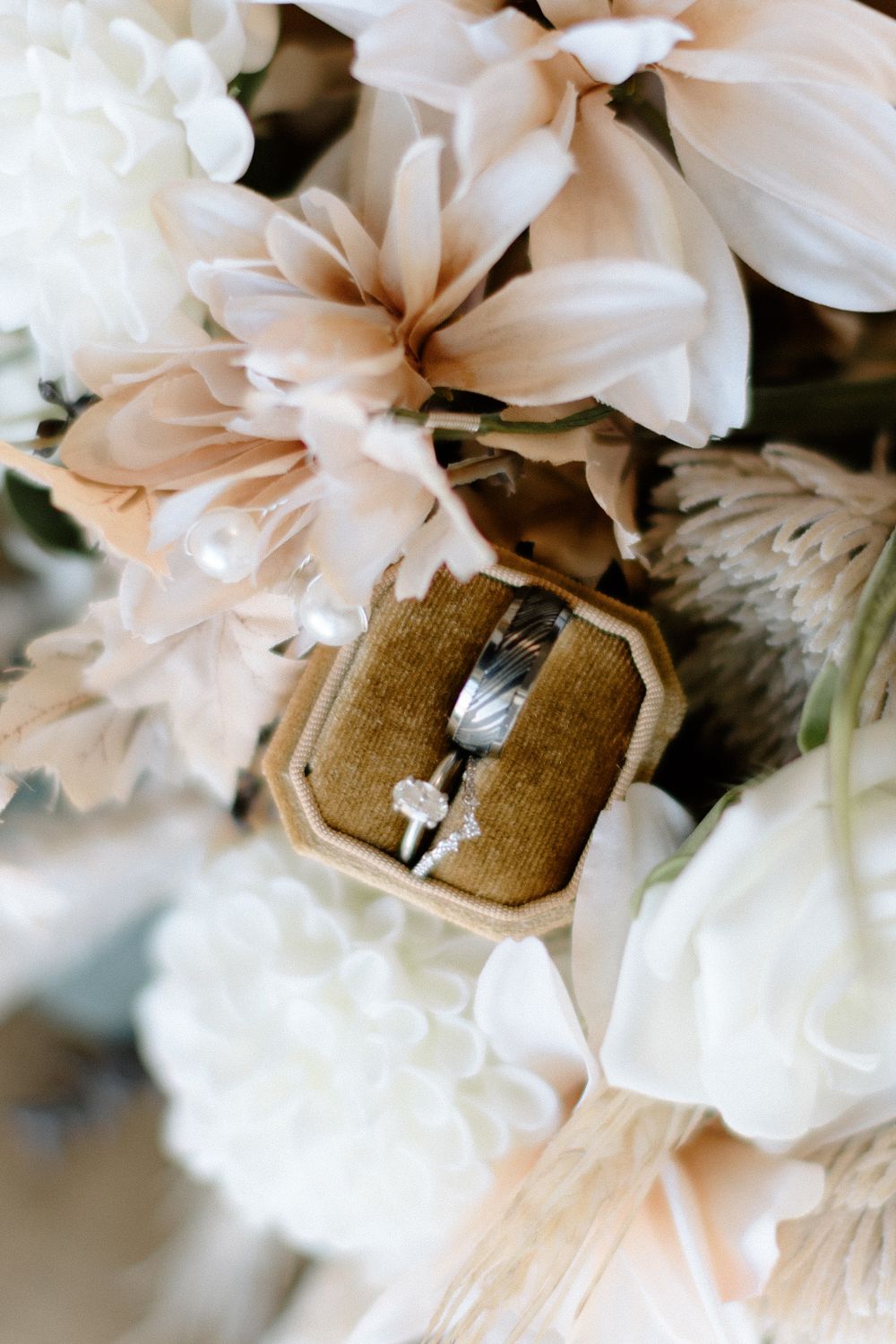 Wedding ring in velvet brown ring box surrounded by soft peach and white flowers in elegant arrangement.