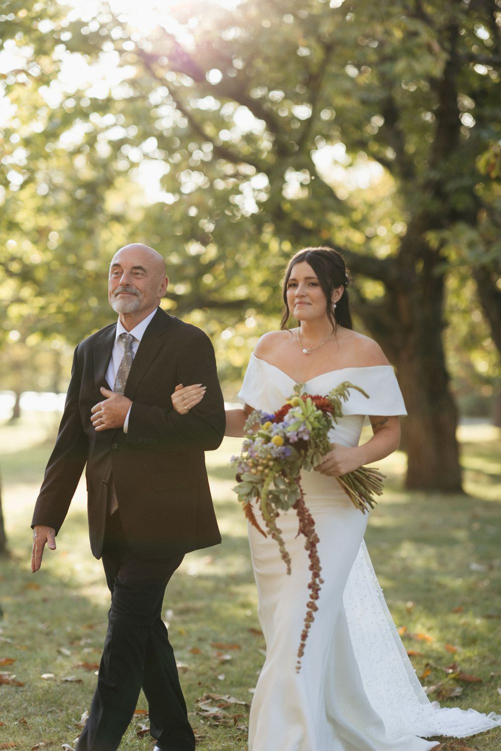 A bride in a white off-shoulder gown walks arm-in-arm during a sunlit outdoor wedding ceremony with a beautiful bouquet.