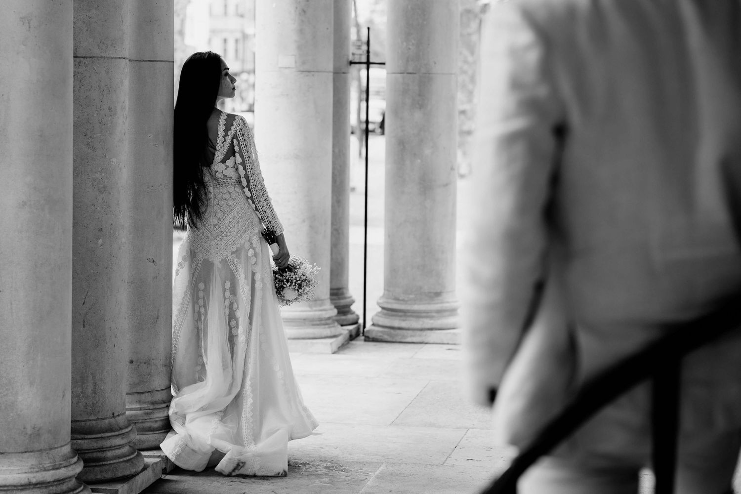 Black and white photo of a bride in wedding dress standing between classical columns as groom looks on in foreground.