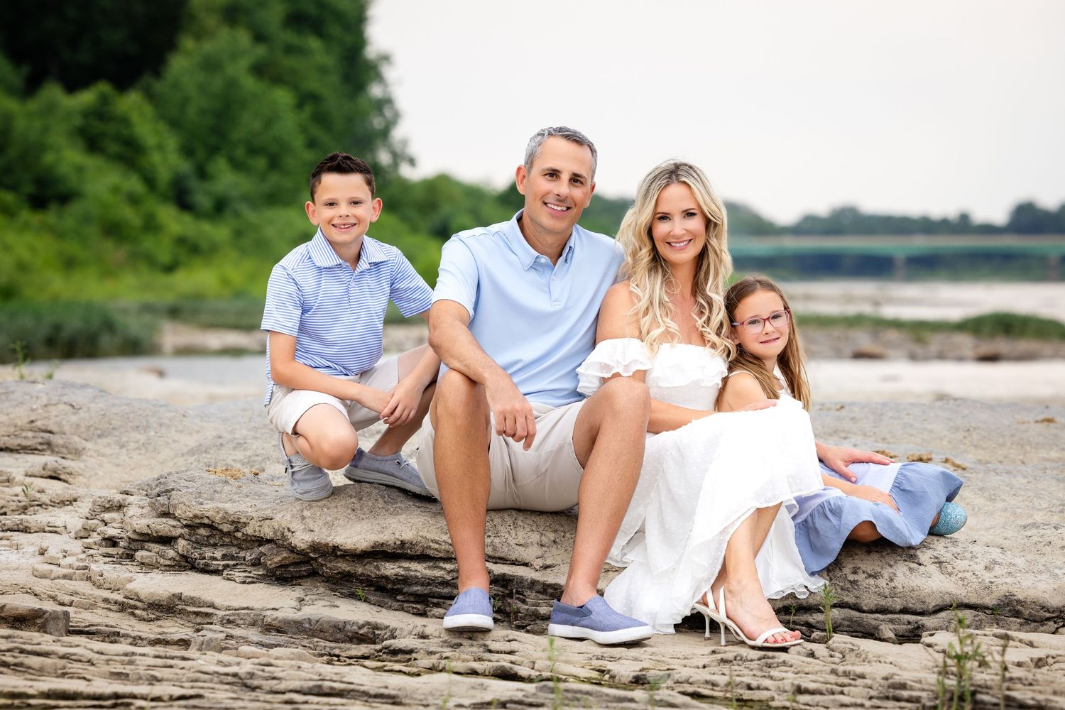 A family in coordinated blue and white outfits sits together on a rocky beach during a summer photo session.