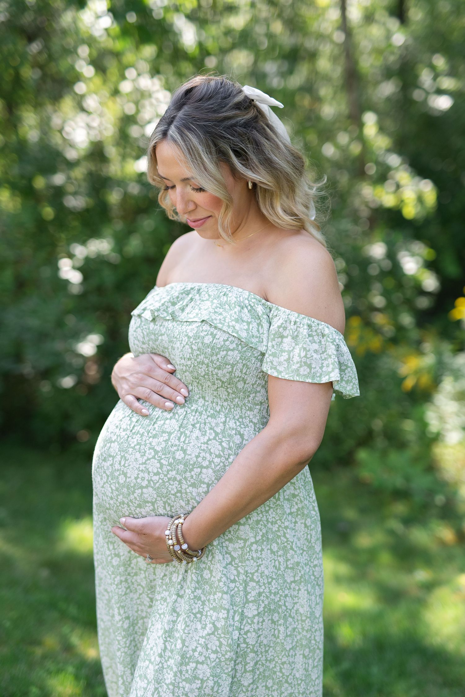 Expectant mother in off-shoulder floral dress poses outdoors while gently touching her baby bump.