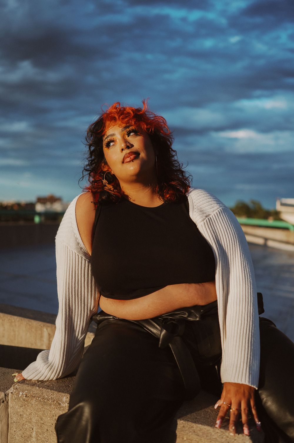 Woman in black tank top and white cardigan poses outdoors against dramatic cloudy sky at sunset.