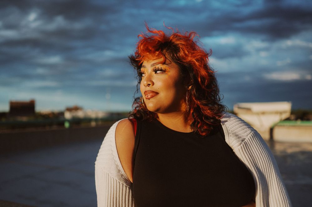 A person with red hair poses outdoors at sunset wearing a white sweater while dramatic clouds loom in the sky.