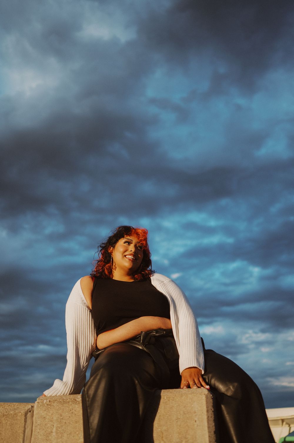 Person in a black top leans back against concrete blocks at sunset with dramatic cloudy sky overhead.