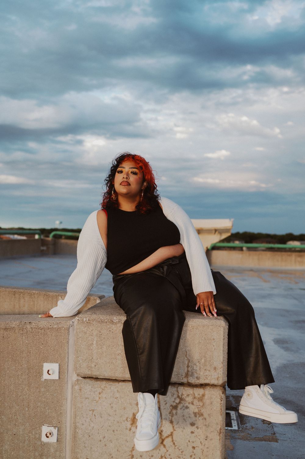 Casual portrait on concrete wall at sunset wearing black outfit with white cardigan and sneakers.