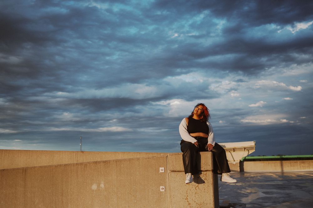Dramatic moody clouds fill the sky at sunset over a lone figure sitting contemplatively on a bench by the ocean.