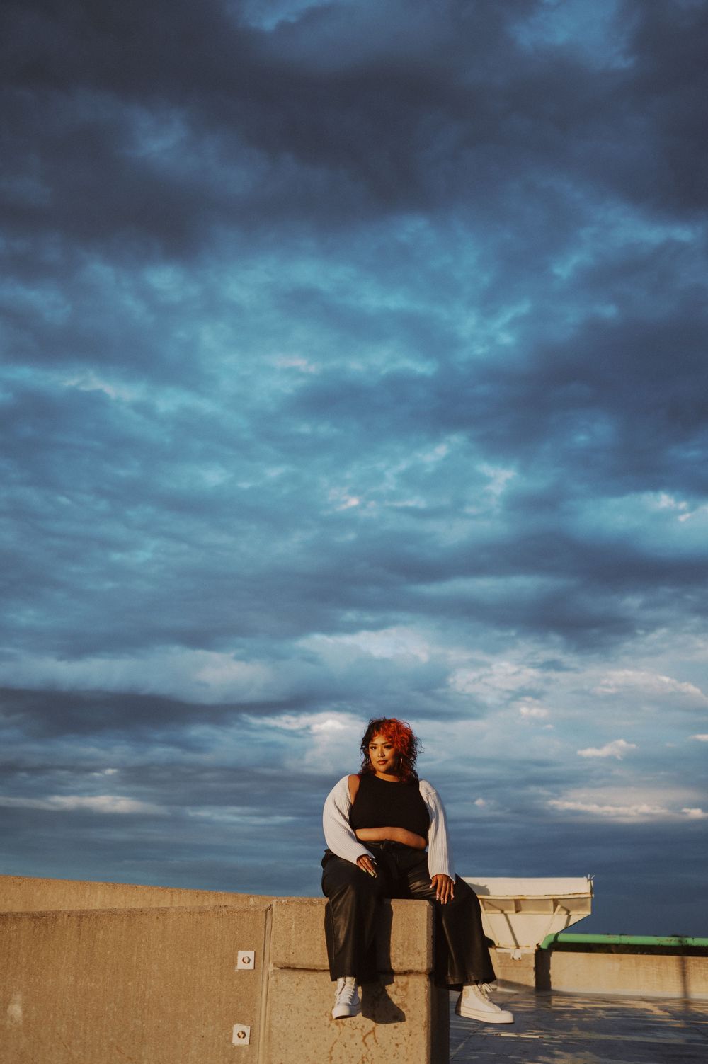 Figure sits on concrete ledge against dramatic stormy blue sky at dusk.