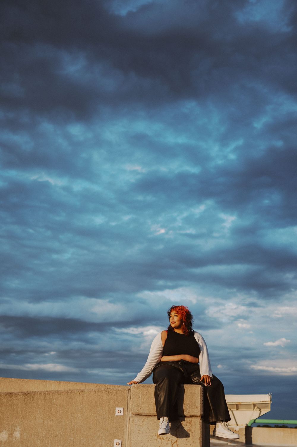 Person sits contemplatively on a wall against dramatic blue stormy sky during sunset.
