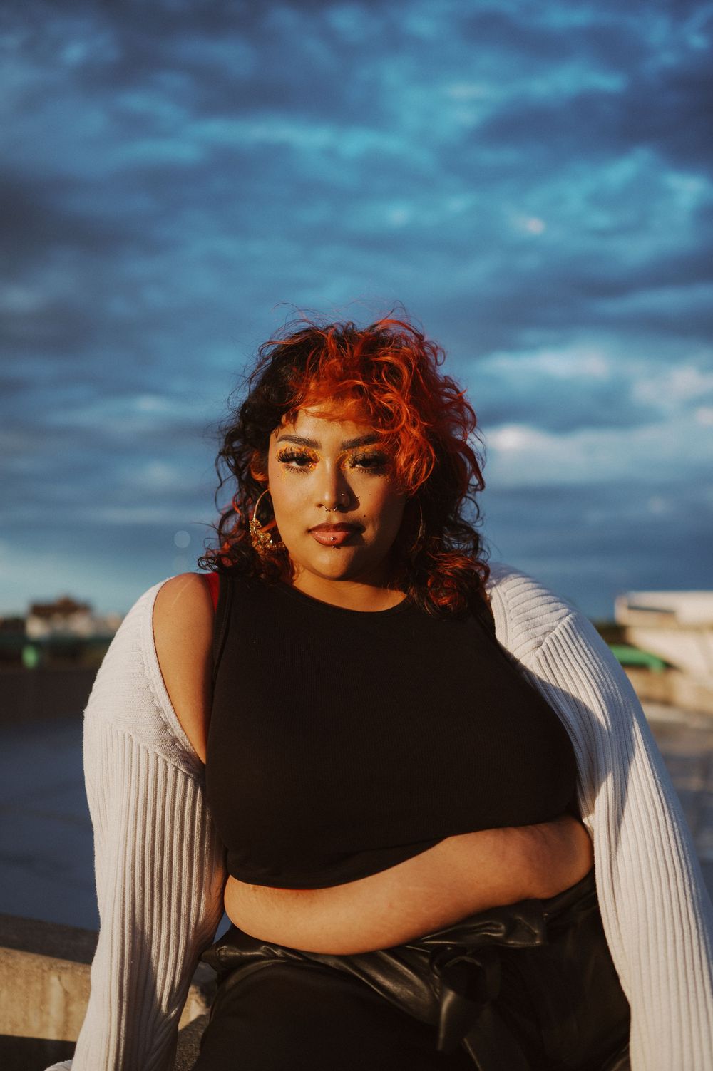 A person with red curly hair poses against dramatic stormy clouds at sunset while wearing black and white clothing.