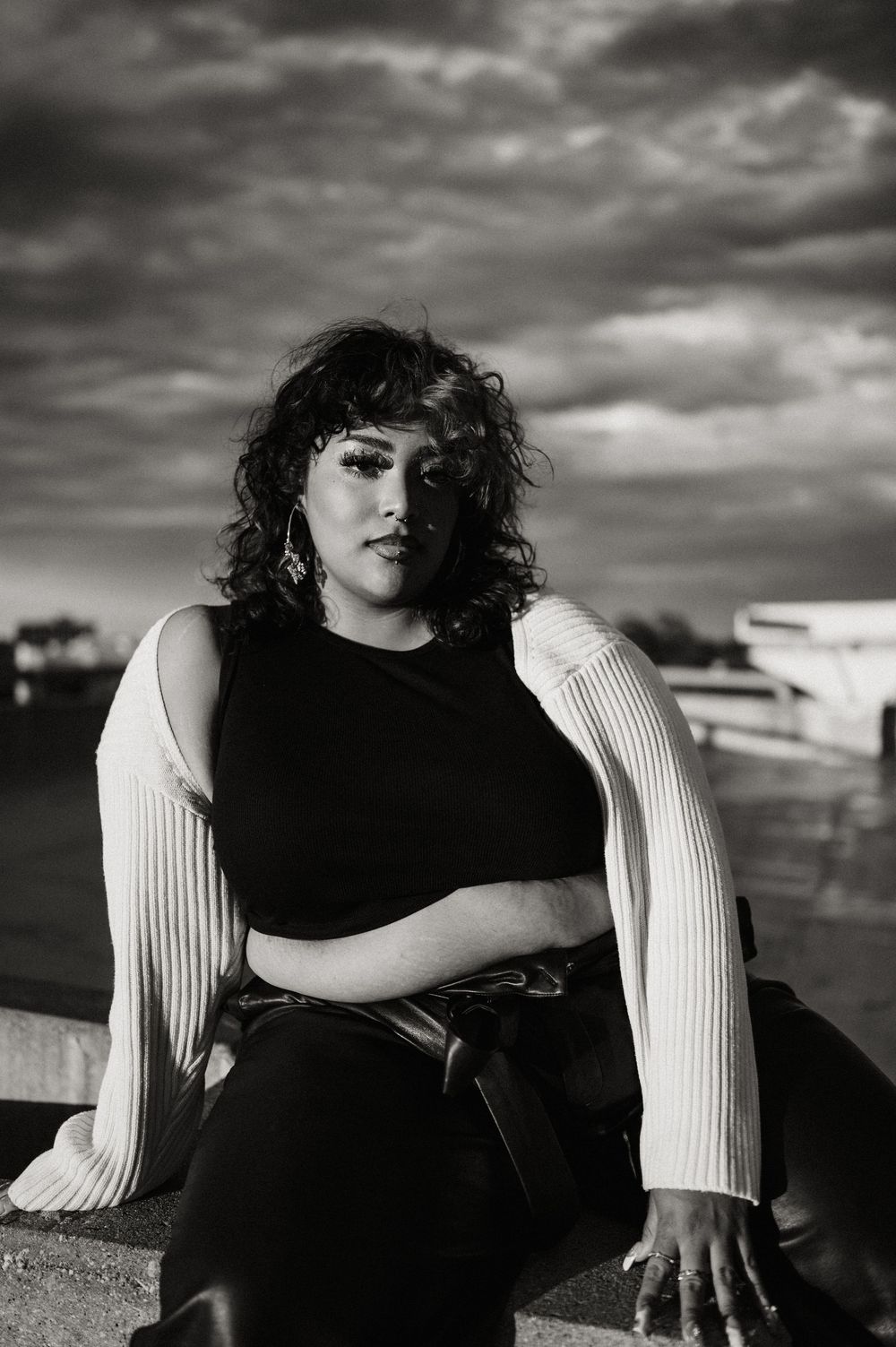 Dramatic black and white portrait under moody clouds featuring curly hair and white knit sleeves against a cloudy sky.