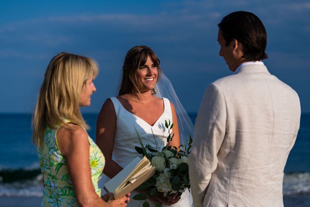A beachside wedding ceremony takes place with three people gathered by the ocean against a blue sky backdrop.