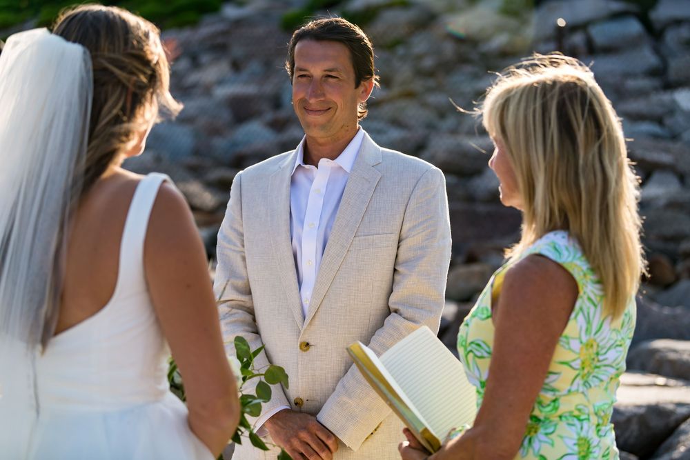 Wedding party dressed in light beige and green floral outfits gather for outdoor photos on rocky terrain near water.