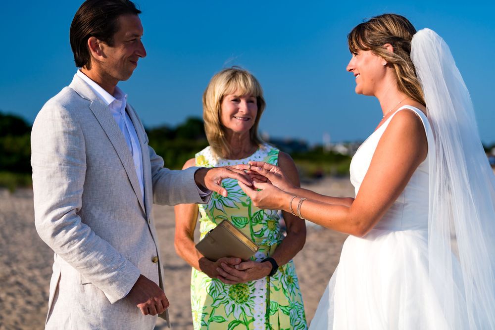 A beach wedding ceremony with an officiant in green floral dress and couple exchanging vows against blue sky backdrop.