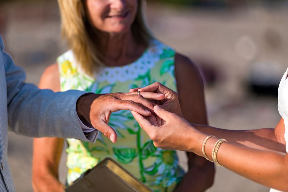 Close up of hands exchanging wedding rings during outdoor ceremony with officiant in floral dress.